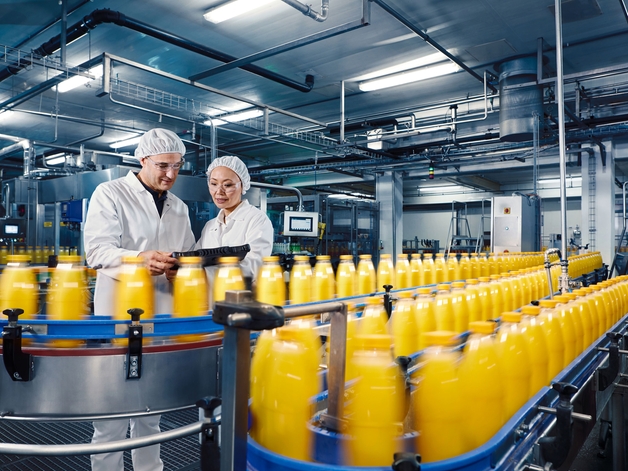 Kaksi työntekijää juomatehtaalla valvomassa appelsiinimehun täyttöprosessia.